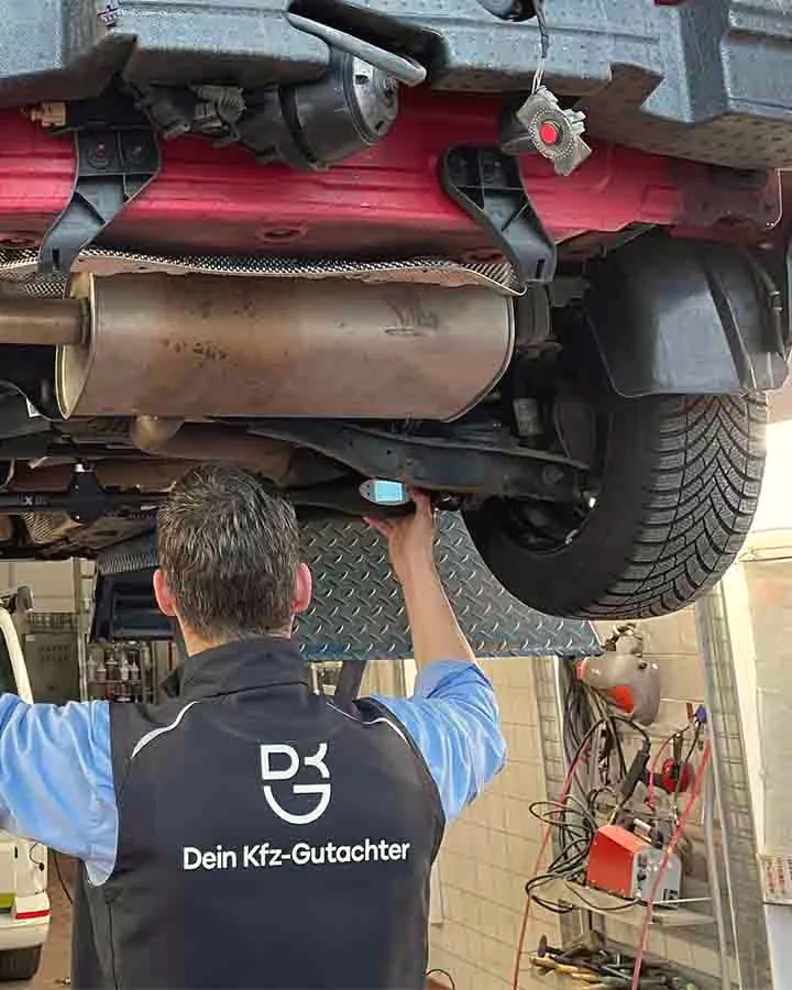 Ein Mann in Weste mit dem Logo „Dein Kfz-Gutachter“ steht von hinten unter einem angehobenen Unfallauto